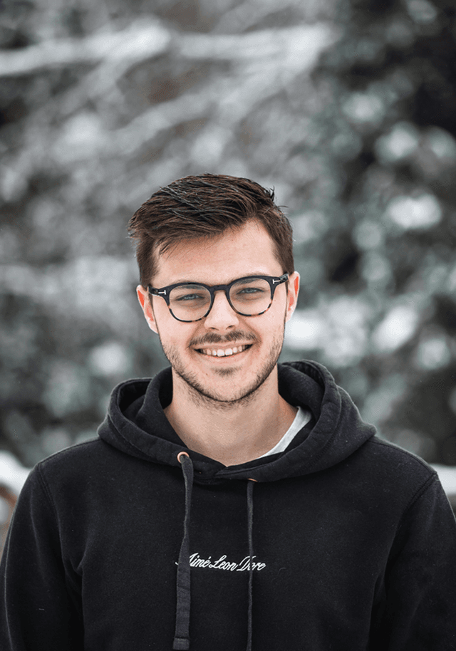 A young man with short brown hair and glasses, smiling in a snowy outdoor setting. He is wearing a black hoodie and has a friendly expression. The background is blurred with snow and trees.
