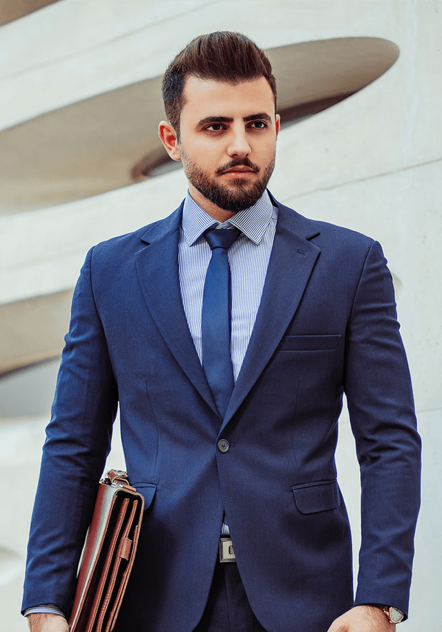A well-dressed man in a navy suit and tie, holding a brown briefcase. He has a well-groomed beard and is standing against a modern architectural background. His expression is serious and confident, with direct eye contact.